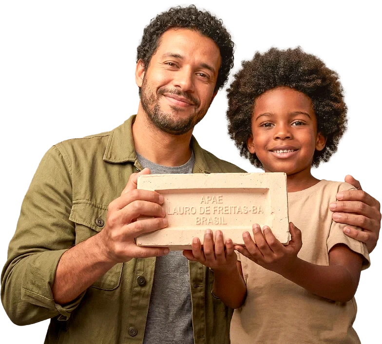 Pai e filho sorrindo, segurando juntos um tijolo com a marca da APAE Lauro de Freitas - BA